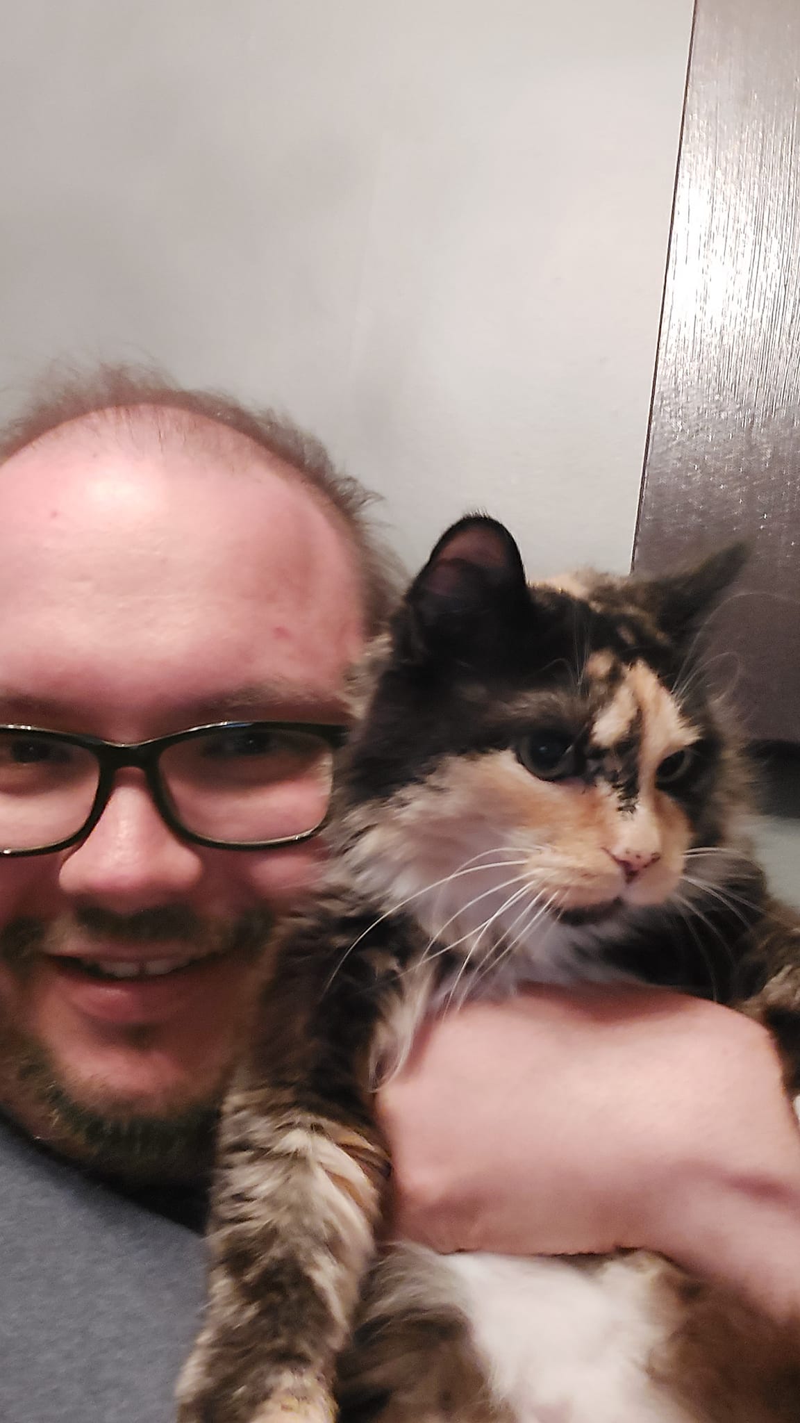 A man sits holding a calico cat, she appears to accept the situation